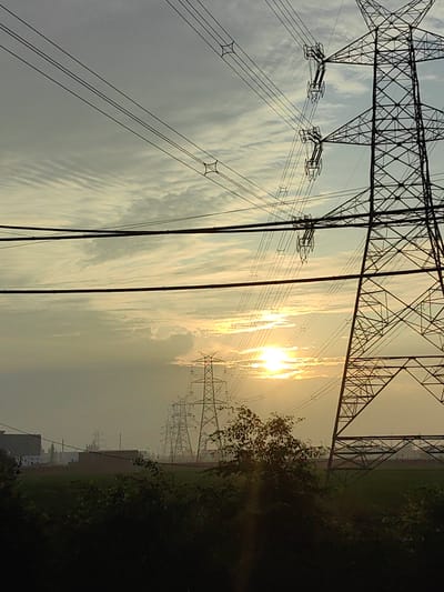 The image depicts a sunset scene with multiple electrical transmission towers stretching across the landscape. The sky is filled with wispy clouds, illuminated by the setting sun, which appears as a bright orb behind a band of clouds. Power lines are strung between the towers, creating a network of lines against the sky. In the foreground, there is dark foliage, suggesting trees or bushes. In the middle ground, there are some buildings, possibly industrial or residential, and a flat, green field. In the background, more transmission towers are visible, receding into the hazy distance. The overall impression is of an industrial or semi-rural area at dusk. The location is identified as Gharaunda, India. There are no people or animals visible in the image.