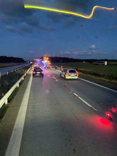 The image captures a nighttime scene on a highway, likely near Långås, Sweden, following an incident. A police car, identifiable by its yellow and blue markings, is parked on the right side of the road, its emergency lights flashing. Further ahead, a large truck is stopped, also with its hazard lights activated. A black passenger car is positioned behind the truck. Several cones and flashing red lights are placed on the road to mark the scene. The sky is dark blue with scattered clouds, and a wind turbine is visible in the distance, suggesting a rural or semi-rural setting. The overall impression is one of an active emergency response on a highway.