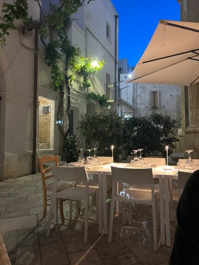 The image depicts an outdoor dining scene at night in Polignano a Mare, Italy. Tables are arranged linearly, covered in white cloths, and set with multiple wine glasses. Seating consists of white plastic chairs, some with wooden accents. Lighting is provided by small lamps on the tables and external building lights. The buildings surrounding the tables are made of white stone and feature traditional architectural elements like windows, balconies, and stone masonry. Vines and greenery are growing along the walls. The sky above is a deep blue, and a large white canopy provides partial cover over the dining area. The ground is paved with light-colored stones. No people are clearly visible at the tables, but a person's arm is visible in the top right corner.