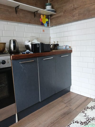 The image shows a corner of a kitchen with white subway tile backsplash and a wooden countertop. There are dark gray cabinets below the countertop with silver handles. To the left, part of a silver oven is visible. On the countertop, there is a silver electric kettle, a bowl with dishes, a black object with a logo that reads "24", a bottle of water, and two decorative soap dispensers. A colorful flower-shaped decoration is attached to the wall above the countertop, near a shelf bracket. A small orange asterisk-shaped decoration is also on the wall. The floor is made of wood-patterned laminate. The lighting suggests it is daytime. The location is not specified in the image.