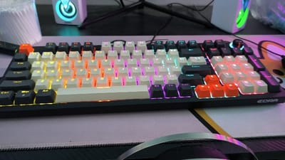 The image shows a black mechanical keyboard with white keys illuminated by multicolor LEDs. The keyboard is positioned on a pink desk mat with a silver headset partially visible in the bottom right corner. The scene is illuminated by artificial lighting. The keyboard is labeled "EDRA" and features standard keys including "Shift", "Ctrl", "Win", and "Tab". The keyboard is illuminated in a rainbow color pattern, with yellow, orange, red, pink, purple, and blue LEDs illuminating the keys. The keyboard appears to be new and unused. The coordinates provided (10.713, 106.45) correspond to a location in Thành phố Hồ Chí Minh, Vietnam, but this location is not visible in the image. The image is a close-up of the keyboard and does not provide any information about the surrounding environment or the person who took the image.