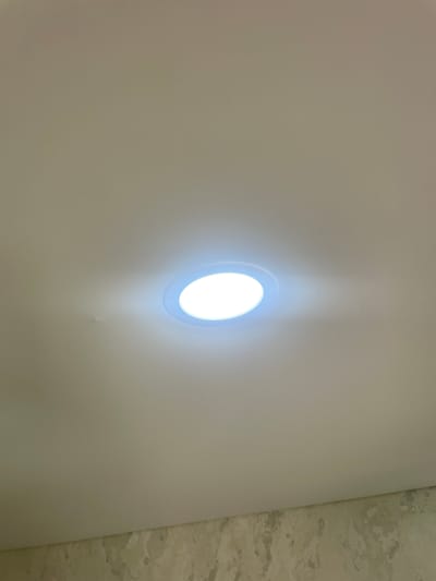 The image shows a close-up of a textured, off-white ceiling with a recessed LED light fixture. The light is on and emits a bright, cool-toned illumination that creates a soft glow and casts subtle shadows on the textured surface. The ceiling appears to be a flat panel, possibly made of drywall or a similar material. To the right of the main ceiling area, a section of what appears to be a patterned wallpaper or a different textured surface is visible, with irregular splotches of light and darker green or gray tones. The overall impression is of an indoor setting, possibly a room or hallway, with a modern lighting element. There is no visible text or any indication of the time of day or weather conditions. The scene is devoid of people or any other objects, focusing solely on the lighting and ceiling textures.