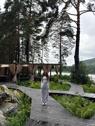 A young woman with long, curly brown hair stands on a wooden boardwalk surrounded by lush green ferns and tall pine trees. She is wearing a white cropped top and gray sweatpants. The boardwalk curves through the landscape, leading towards a covered outdoor seating area with tables and chairs. In the background, a river or lake is visible, with hills and trees on the opposite bank. The sky is overcast. The location is identified as Nizhnekayancha, Russia.