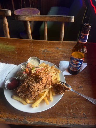On a weathered wooden table, likely at a restaurant in Santa Cruz de Yojoa, Honduras, sits a plate of fried chicken, fries, and coleslaw with two small metal bowls of dipping sauces. A bottle of Salva Vida beer lies on its side on a white napkin. A fork pierces a piece of the fried chicken. Beside the table is an aged wooden chair with a round, painted seat.