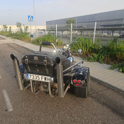 A black three-wheeled motorcycle is parked on a paved surface next to a sidewalk in Illescas, Spain. The motorcycle has two large chrome exhaust pipes on each side and a black leather storage compartment with studs and buckles. The license plate reads "7335 FWH". In the background, a chain-link fence separates the motorcycle from a large, modern building with a gray facade and a row of lights. Green bushes and trees are visible along the fence line. A pedestrian crossing sign is also present near the building. The sky is clear and blue, indicating daytime.