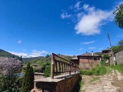 The image captures a sunlit scene in Kovachevitsa, Bulgaria. In the foreground, a rustic wooden fence tops a stone wall. Behind the fence, a stone and brick building with a slate roof stands. The path to the right is made of rough, uneven stone, flanked by a stone wall and a telephone pole. In the distance, wooded hills rise under a bright blue sky dotted with scattered clouds. A flowering tree with purple blooms adds a splash of color to the left side of the image.