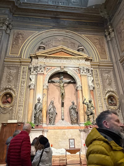 This image depicts the main altar inside the Cattedrale di San Giorgio Martire (Ferrara Cathedral) in Ferrara, Italy.
The scene is dominated by a grand altar, centrally featuring a large, gilded bronze crucifix with the figure of Christ. Below the crucifix, four life-sized bronze statues stand on individual pedestals: from left to right, a robed bishop holding a staff, a robed female figure, a robed male figure, and an armored male figure wielding a spear. These statues are positioned within an elaborate architectural framework comprising marble columns, pilasters, and intricate classical carvings with gold accents. Above the crucifix, a semi-circular arch contains a Latin inscription: "PRO DEO, REGE, ET LEGE, ET CHRISTO". Further decorative elements, including sculpted angels and cherubs, adorn the walls above and to the sides of the main structure.
To the left and right of the central altar, integrated into the wall decoration, are two circular gilded frames each containing a red-toned bust. The frame on the left is labeled "S. THOMAS". Below the main statues, a white marble base serves as an altar or tomb, with a visible inscription plate mentioning "BONAVIA ROLLI ARCHIEPISCOPUS FERRARIAE PASTOR ET DEFENS". A small wooden table on the right of this base holds a framed image of a crucifix.
In the foreground, several individuals observe the altar. On the left, a balding man in a red puffer jacket stands with his back mostly to the camera, looking towards the altar. Adjacent to him, a person in a dark jacket is partially visible. Behind them, two women, one with dark hair in a light coat and another in a black beanie, are also facing the altar. On the right, a man with a dark beard, wearing a mustard yellow puffer jacket, is seen in profile, looking towards the right side of the frame, partially obscuring the lower right portion of the altar. The overall environment is dimly lit, characteristic of a large cathedral interior.
