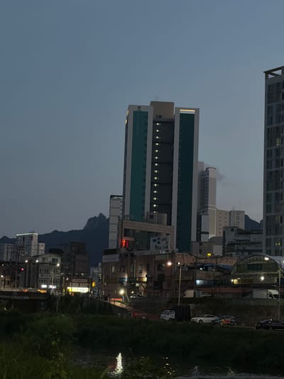 The image depicts a twilight scene in Uijeongbu-si, South Korea, featuring a cityscape with several tall buildings, including a prominent teal and white apartment complex with visible lit windows. In the background, a distinctive mountain range with jagged peaks is silhouetted against the darkening sky. The foreground shows a river with reflected lights, bordered by lush green vegetation along its banks. A road runs alongside the river, with a few vehicles, including a white car and a smaller vehicle with red and blue decals, visible. Streetlights and building lights illuminate parts of the urban environment. The atmosphere suggests a transition from day to night in a city.