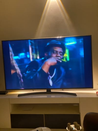 A man with headphones and a chain is displayed on a television screen. The screen has a dark blue hue. He is holding something up to his mouth. In the background, there are blurred blues and greens. A light beam is visible on the wall to the left of the TV. To the right of the TV is a cabinet with dark and light panels.