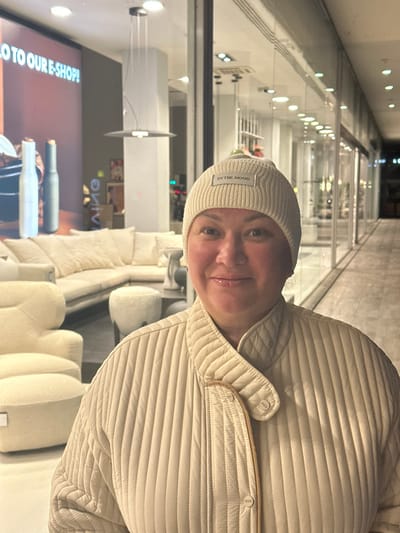 A woman is featured prominently in a well-lit indoor setting, likely a commercial space such as a furniture store or showroom in Engomi Lefkoşa, Cyprus. She is seen from the chest up, looking directly at the camera with a gentle, slightly smiling expression.

She is wearing a cream-colored, ribbed knit beanie on her head, which has a small rectangular label on the front that reads "IN THE MOOD." Her attire also includes a matching cream or off-white quilted jacket with snap buttons, suggesting a comfortable and casual style.

The background reveals a modern interior with various light-colored, plush furniture items, including sofas and chairs, arranged as if in a display. The ceiling features multiple recessed lights, and reflections on glass partitions lining a corridor or pathway suggest a spacious and open layout. On the far left of the image, a large screen or display is visible, partly obscured. It shows text that reads "...TO OUR E-SHOP!" and features images of product bottles, possibly for beverages like wine or spirits. The overall lighting is artificial and bright, suggesting an indoor evening or night scene, or simply a brightly lit retail environment.