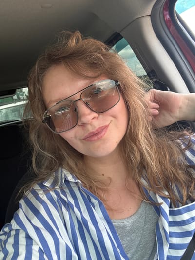 A young woman with long, wavy blonde hair, wearing sunglasses and a blue and white striped shirt over a grey t-shirt, is taking a selfie in a car. The interior of the car is visible, including the passenger-side door frame and part of the dashboard. The woman is touching her hair with her right hand. The scene is likely taking place in the city of Kachimir, Russia, as indicated by the prompt.