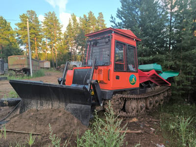 In the city of Gremuchiy, Russia, a tracked tractor, painted bright orange and black, is positioned on a dirt path. The tractor is equipped with a large black front blade, which holds a pile of brown soil. The machine has a reinforced cab with black-grated windows. A green attachment is visible on the rear right side of the tractor. The background features a dense forest of tall pine trees and a clear sky with some clouds. To the left of the tractor, an old, weathered green trailer is parked, and further left, a utility pole stands. The ground is covered with dirt, grass, and some scattered debris. The overall scene depicts an outdoor industrial or agricultural setting.