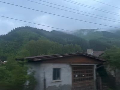 The image shows a gray, overcast scene with mountains in the background partially obscured by fog. The lower portion of the frame features a building with a brown-trimmed roof, a small window, and a garage door. Overhead power lines are visible against the sky. The location is possibly near Lukovo, Bulgaria, based on the prompt. The scene appears to be taken from a moving vehicle.