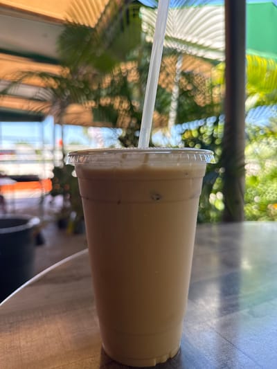 A close-up, slightly sideways image shows a clear plastic cup with a straw, filled with a light brown beverage, likely an iced coffee or similar drink. The cup has a ridged lid and condensation is visible on the side.
In the background, there is a blurred outdoor scene, featuring a combination of green foliage and architectural elements. The light and color suggest a sunny day. The blurred vegetation appears to be palm trees, giving a tropical or subtropical impression.
Based on the information provided about the location being in Davie, Florida, the vegetation is consistent with the climate and typical landscaping of that area. The beverage is consistent with being purchased from a local coffee shop in the outdoor dining area.
