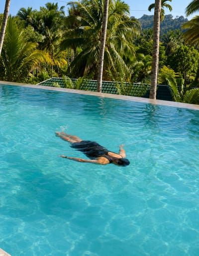 A person wearing a black swimsuit swims in a turquoise infinity pool. The pool is bordered by a concrete edge and green tiled roofing. In the background, lush green palm trees and tropical vegetation cover rolling hills under a clear blue sky. The person is shown from above, swimming with their face submerged in the water.