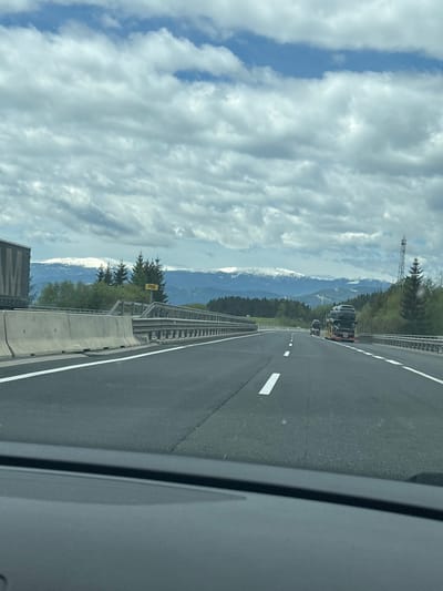 The image depicts a view from inside a vehicle driving on a highway. The road has multiple lanes and a concrete barrier. Mountains are visible in the distance, partially covered with snow. Clouds dominate the sky. A truck is in the lane ahead. The location is identified as Unterauerling, Austria. There is no indication of any specific interaction. The environment consists of a highway surrounded by mountainous terrain. A highway sign is visible saying P08.