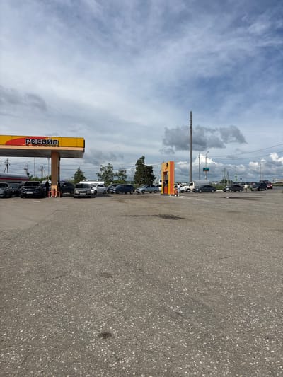 This is a ground-level, full-shot photograph of a gas station in the city of Kleschevka, Russia, under a cloudy sky. The gas station canopy, emblazoned with the logo "РОСОЙЛ" in red and yellow, is prominently featured on the left side of the frame. Several cars are lined up at the gas pumps, suggesting a queue. To the right of the canopy, a bright orange and yellow gas pump structure stands, with two individuals near it. One person appears to be a customer, while the other, dressed in a light-colored shirt, is operating the pump. A white truck is parked behind the pump structure. Further in the background, more vehicles are visible, including cars and possibly a truck, on a roadway that appears to lead out of the gas station area. Trees and utility poles are scattered throughout the background. The foreground is dominated by a paved asphalt surface, showing signs of wear and tear with some darker patches that may indicate oil stains. The overall impression is a typical operational scene at a gas station.