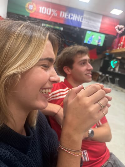 This image depicts an indoor scene with two young adults in Décines-Charpieu, France. The location is identified by a banner in the background displaying "100% DECINES" overlaid on a combination of the Portuguese and French flags.
In the foreground, a light-skinned female with shoulder-length blonde hair is shown in profile, facing right. She is smiling broadly, revealing upper and lower teeth, and her eyes are crinkled. She wears a dark blue knitted sweater. Her hands are clasped together; her right hand shows a gold ring on her ring finger and another thin ring on her middle finger. Her left wrist features multiple bracelets, including a brown band, a red and white braided cord, and a thin gold chain.
Behind her and to the right, a light-skinned male with short brown hair is also visible in profile, facing right and looking in the same direction as the female. He wears a red polo shirt with white horizontal stripes. His right arm is partially visible, appearing to rest near the female. He wears a silver-colored wristwatch on his left wrist and a thin red string bracelet on his right wrist.
The background includes a flat-screen television displaying a blurred image of what appears to be a soccer match with two players. To the right of the television, red decorative items, including a heart-shaped ornament, are visible. A blurred green light fixture is also present further right. The floor is light-colored. Both individuals appear to be focused on an activity or view to their right or on the television.