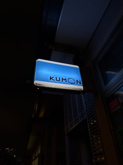 This is a nighttime, medium-shot, eye-level photograph of a Kumon educational center's illuminated sign on a building in Chiba, Japan. The sign, a horizontally oriented rectangle with rounded corners, is blue and white and displays the Kumon logo and text. The logo, featuring a smiling face within a circle, is to the right of the text "KUMON". The sign is mounted on the exterior of a building.

To the right of the sign, a window reflects some of the surrounding environment, and a red sticker with Japanese text and the word "SECOM" is visible on the building's wall. Below the sign, a dark, horizontally-slatted structure is prominent. In the background, a dark, modern building with many illuminated windows is visible in the distance, suggesting an urban setting. The overall lighting is dim, with the Kumon sign being the primary light source. The photograph is taken from a slightly upward angle.