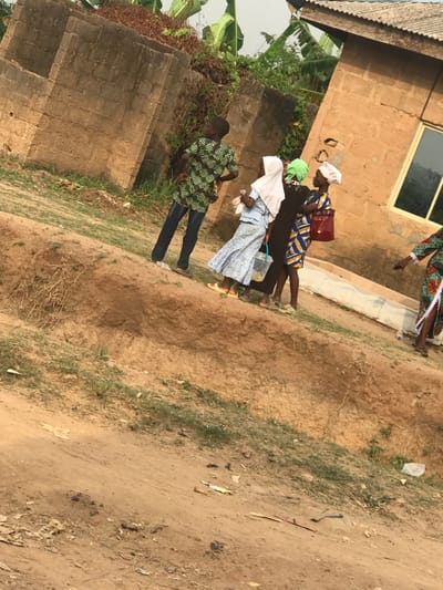 Four people walk along a path adjacent to a building. The person nearest the viewer is a child in jeans and a patterned green shirt. Behind them, three women walk in a line. They wear long dresses, two wear head coverings and the third wears a headscarf. One woman carries a red bag. The path is dirt and grassy. The building is brick with a corrugated metal roof and a window.