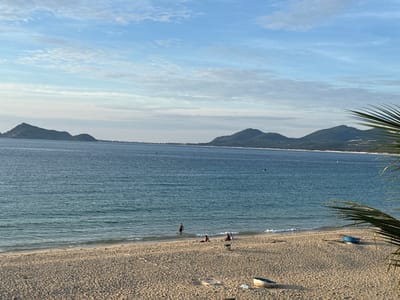 This image captures a scenic view of a tropical beach in Song Cau, Vietnam. The foreground is dominated by a sandy beach with scattered debris and two small boats, one blue and one white, resting on the shore. In the middle ground, the calm, blue ocean meets the shoreline, with gentle waves lapping at the sand. Several people are visible on the beach and in the shallow water. One person stands in the water, while others are seated or lying on the sand. The background features a distant coastline with lush, green hills and mountains under a bright blue sky dotted with white clouds. A palm frond partially frames the right side of the image. The overall atmosphere is one of tranquility and a leisurely day at the beach.