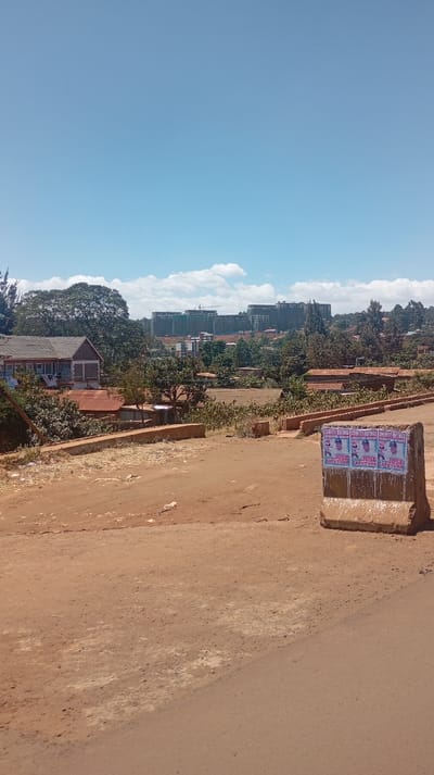 An outdoor scene features a dirt road in the foreground. Behind it, buildings are partially obscured by trees. In the distance, larger buildings under construction rise on a hill. A concrete barrier with posters is visible on the right. The sky is clear blue with a few scattered clouds. The overall scene suggests a rural or developing area undergoing construction.