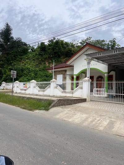 This is a wide, eye-level shot of a house in Rantau Pauh, Indonesia, on a cloudy day. The house is white with green accents around the porch and red doors. It has a brown, gabled roof and a metal carport. The house is surrounded by a white, ornamental fence. A paved driveway leads to the house, and a street runs in front of it. Lush green trees and foliage rise up behind the house on a gentle slope. A street sign is visible on the left side of the frame, and a sign advertising a house for sale is attached to the fence. There are no people visible in the image. The overall impression is one of a residential street on a slightly overcast day.