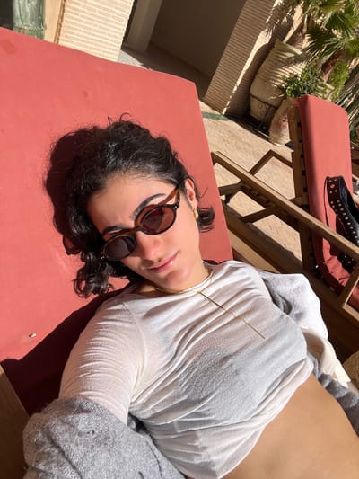 A female individual is depicted reclining on a red fabric chaise lounge with a wooden frame, outdoors under bright sunlight. She has dark, curly hair and is wearing tortoiseshell-framed oval sunglasses, small gold-colored stud earrings, and a thin gold-colored necklace featuring a vertical bar pendant. Her attire consists of a white, long-sleeved, somewhat sheer top, with a grey knitted cardigan draped over her lower torso and legs. Her right arm is visible, with the hand out of frame.

The setting is an outdoor courtyard or patio, identified as being in Rissani, Morocco. The ground surface is paved with light-colored tiles. To the left and in the background, light-colored brick or stone architectural structures are visible, consistent with Moroccan design. To the right of the primary subject, another identical red chaise lounge is present, on which a pair of black, studded, flat shoes are resting. Further in the background on the right, large terra cotta-colored ceramic pots and green foliage, including palm fronds, are visible. Strong shadows are cast, indicating direct overhead sunlight. The individual is positioned looking towards the camera, with a slight smile.