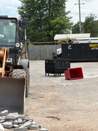 In Norwood, USA, a construction site features an orange and black front-end loader in the foreground. To the left, a pile of broken gray paving stones rests against the loader's bucket. Behind the loader, a black plastic crate with wheels sits on the gravel lot. A red plastic bin lies on its side next to the crate. In the background, a large black dumpster is positioned near a wooden fence. A white van is partially visible next to the dumpster, which has "WESTERN WASTE" and a phone number printed on its side. Overhead power lines stretch across a cloudy sky, and lush green trees frame the scene.