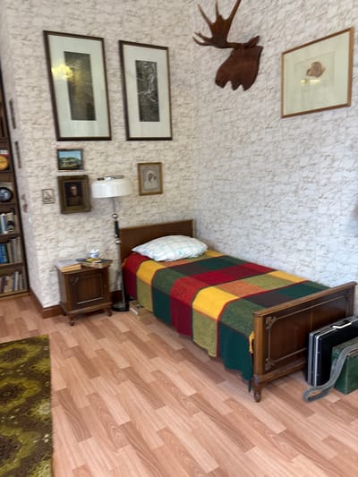 The image depicts a bedroom scene with a single bed. The bed is covered with a colorful, plaid blanket and a white pillow. To the left of the bed is a wooden nightstand with a lamp and some books on top. The walls are covered in patterned wallpaper. Above the nightstand, there are several framed pictures on the wall. To the right of the bed, a moose head mount is visible on the wall, along with another framed picture. The floor is made of light-colored laminate. A small portion of a patterned rug is visible in the lower left corner. The room appears to be a bedroom, possibly in a rustic or vintage setting. There are no people visible in the image. The location is identified as Овсянка, Russia.