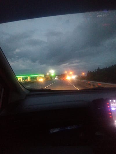 The image depicts a view from inside a vehicle traveling on a highway at dusk. In the distance, a brightly lit gas station with green illuminated canopies is visible. Several vehicles are present on the highway, some with their headlights on, illuminating the wet road surface, suggesting recent rain. The sky is overcast with dark clouds. The interior of the car, including the dashboard and a portion of the windshield, is visible in the foreground. The dashboard shows a digital display with illuminated buttons. The scene is consistent with a highway scene in or around Vasilyevka, Russia, given the time of day and the presence of a service area.