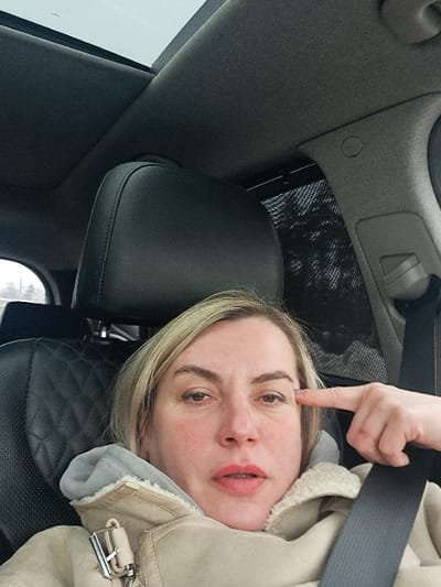 The image shows an adult female seated in the driver's seat of a vehicle, in Obukhovo, Russia. She has blonde hair and is wearing a beige winter coat with a faux fur lining and a visible metallic buckle. A black seatbelt is positioned across her chest and lap. Her right hand is raised, with the index finger pointing towards her right temple. She is looking directly at the camera. The vehicle's interior includes a dark headliner, a black leather seat with a quilted pattern on the seatback, and a panoramic sunroof overhead revealing an overcast sky. A dark mesh sunshade is visible on the passenger side window, partially obscuring blurred outdoor vegetation.