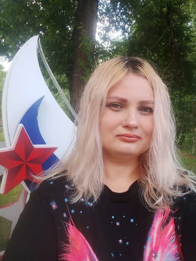 A medium shot shows a woman with bleached blonde hair and light skin, wearing a black t-shirt with a pink and blue feather design. She has makeup on, including foundation, blush, eyeshadow, and lipstick. She is standing outdoors in front of a large, white, crescent-shaped sculpture with a red star at its base, against a backdrop of green trees and a brown tree trunk. The scene appears to be located in the city of Изыхские Копи, Russia.