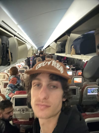 The image depicts the interior of a commercial aircraft cabin, observed from a forward-facing perspective, taken inside an aircraft at Charles de Gaulle Airport (CDG) in Tremblay-en-France, France. In the immediate foreground, a male individual with medium-length dark wavy hair and a light brown baseball cap, adorned with white embroidered text or a logo, looks directly at the camera. He is wearing a dark garment.
Behind the foreground subject, the aircraft aisle extends rearward. To the lower left, a man with dark hair and a beard is partially visible, looking downwards and to his left. Further back on the left side of the aisle, a child with reddish-brown hair, wearing a multi-colored jacket (patterns of blue, orange, and white), is seated in an aisle seat, facing away from the camera. Multiple other passengers are visible standing and seated throughout the cabin, populating the aisle and seat rows.
The cabin features standard aircraft seating with individual entertainment screens on the seatbacks; several screens are illuminated, displaying graphical interfaces consistent with flight information or entertainment. Overhead bins are open along both sides of the cabin, revealing various pieces of luggage, including a dark blue suitcase and other bags. The cabin is illuminated by bright, artificial overhead lighting. The scene suggests a state of boarding or deplaning, characterized by a significant number of passengers and accessible overhead storage.