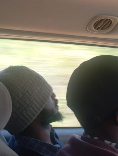 The scene depicts the interior of a moving vehicle in the city of Kwagal, Nigeria, as observed from a rear passenger perspective. Two individuals are visible. On the left, a male subject with a dark beard wears a light-colored, possibly beige or grey, knit beanie pulled down over his eyes, suggesting he is resting or asleep. He is clad in a blue and white checkered button-down shirt. His head is tilted back against a light-colored headrest. To the right, only the back of another individual's head and upper shoulder are visible; this person has short, dark hair and appears to be wearing a dark garment with a patterned or striped collar visible near the shoulder. The vehicle's light-colored ceiling features an oval-shaped air vent. Through the side window, a heavily motion-blurred view of a light green or yellow outdoor environment indicates the vehicle is in motion.