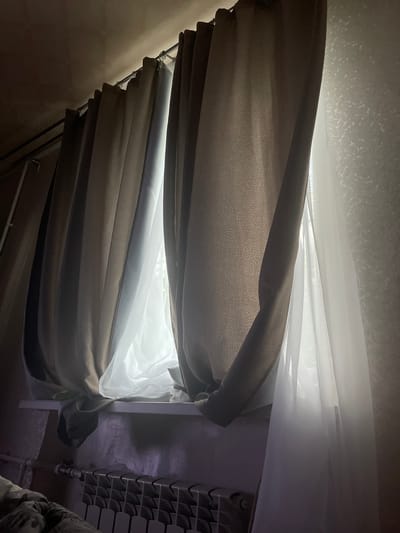 The image captures a dimly lit interior scene focused on a window with layered curtains. Thick, taupe-colored curtains are drawn partially open, revealing sheer white curtains behind them. Sunlight streams through the gap between the taupe curtains and the sheer ones, creating bright highlights and casting shadows. The window sill below is visible, with what appears to be a radiator beneath it. The wall to the right of the window has a textured pattern. The overall impression is one of a private space, possibly a bedroom, in a modest setting. No specific landmarks or architectural details suggest a location within Gukovo, Russia, or any other city.