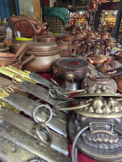 The image depicts a market stall in Gucheng District, China, overflowing with traditional copper and brass cookware and teapots. Numerous teapots of various sizes and designs are stacked and arranged, featuring ornate handles and spouts. Several large copper pots and bowls are also visible, some with intricate patterns. In the foreground, metallic elements with ring handles, possibly part of decorative displays or functional accessories, are laid out. The background hints at a bustling marketplace with shelves of more copper items and stalls. There are no people clearly visible in the frame.
