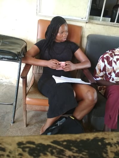 A Black woman with braided hair is seated in a wooden chair with an upholstered seat and back. She is wearing a black short-sleeved t-shirt and a black skirt. She is holding a small pink object in her hands. A stack of papers rests on her lap. To her right, another person is partially visible, wearing a patterned shirt and burgundy pants. A dark bag is on the floor near her feet. The background shows a light-colored wall and a window with bars. The scene appears to be indoors, possibly in a waiting area or meeting room, in Bisichi, Nigeria.