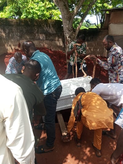 In Oko Town, Nigeria, an outdoor burial scene depicts the interment of a white casket. Approximately eight adult males are present, engaged in various tasks associated with the funeral. A white casket, featuring a textured or speckled surface, is positioned over an open grave, supported by wooden planks. A mound of reddish-brown earth, indicative of excavated soil, is piled behind the grave.
Individuals are observed interacting with the scene:
- A bald man in a light blue short-sleeved shirt and dark jeans is bending towards the casket from the left, seemingly assisting in its placement or lowering.
- To his right, a man in an orange traditional top and trousers is also bending low, reaching towards the ground or the base of the casket.
- In the background, two men wearing patterned short-sleeved shirts are handling long, curved, natural wooden implements, possibly used in the casket's descent.
- Another man in a patterned shirt is visible behind the casket, in the grave area, appearing to be moving soil.
- The immediate environment consists of reddish-brown soil underfoot, a rough concrete wall in the background, and green foliage, including a tree that casts dappled shadows. The event occurs under bright daylight.