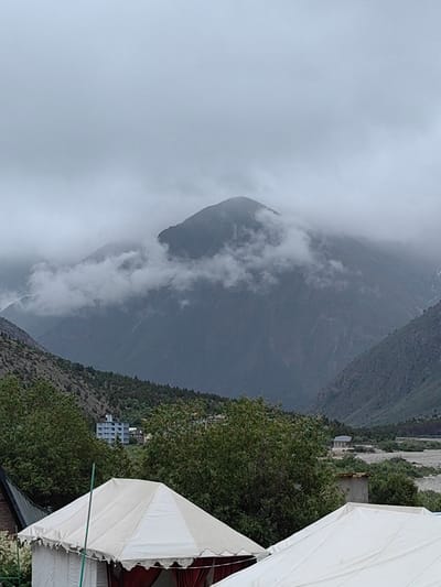 This image captures a view of a mountainous landscape in Jispa, India, under a cloudy sky. In the foreground, two white tents are visible, suggesting a campsite. Behind the tents, lush green trees and vegetation cover the lower slopes of the mountains. A few buildings, including a multi-story structure, are scattered amidst the greenery. The background is dominated by large, imposing mountains shrouded in thick clouds, giving the scene a dramatic and somewhat mysterious atmosphere. A dry riverbed or a wide expanse of rocks is visible in the mid-ground. The overall impression is one of natural beauty and a remote, rugged environment.