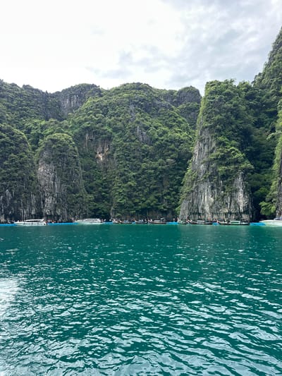 In Haad Yao, Thailand, a body of turquoise water fills the foreground of the image. Gentle waves ripple across the surface, reflecting the overcast sky. In the midground, several longtail boats are moored near a line of blue barriers. Behind the boats, the water meets a shoreline characterized by towering limestone cliffs covered in lush green vegetation. A few people are visible on a floating platform near the boats. The overall scene depicts a tropical coastal landscape.