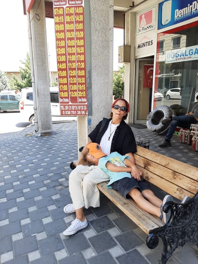 A woman and a child are sitting on a wooden bench in front of a storefront. The woman, wearing a red bandana, sunglasses, and a black shirt over a white t-shirt, is looking towards the camera. The child, wearing an orange cap and a light blue t-shirt, is resting their head on the woman's lap, with their eyes closed. They are seated on a sidewalk paved with dark gray and light gray square tiles. Behind them, a bus schedule is displayed on a sign. The storefront features signs for "Dr. Pimapen," "MINTES," and "DOĞALGA." There is a Turkish flag hanging in the doorway of the store. To the right of the bench, another person is seated on what appears to be a bench or a similar structure. In the background, vehicles, including a white van and a green car, are parked. The scene is set in Nevşehir, Turkey.