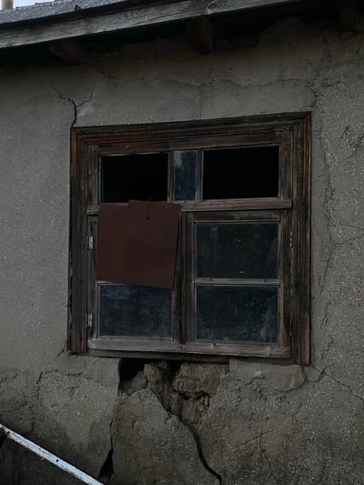 The image shows a close-up of a weathered window on the side of an old building, likely in the village of Cairaclia, Moldova. The building's facade is rough, textured concrete with visible cracks, suggesting disrepair. The wooden window frame is dark and aged, with some peeling paint or finish. The window itself is divided into panes, some of which are intact and dark with reflection, while others are open and black. A square piece of rusty metal is affixed to the inside of the window, covering a portion of one of the panes. Below the window, the concrete is significantly damaged, with large chunks missing and the underlying material exposed. A white metal object, possibly a railing or pipe, is partially visible in the bottom left corner. The overall scene evokes a sense of neglect and abandonment.