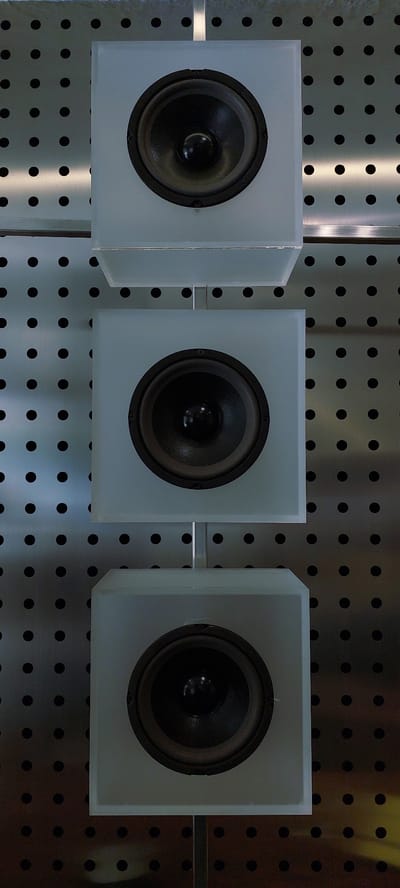 The image displays three speakers mounted vertically in a row. The speakers are encased in frosted white boxes. The background is a metallic surface with a pattern of evenly spaced black dots, suggesting a perforated metal sheet. The lighting creates a highlight that runs vertically down the right side of the image, illuminating the polka dot pattern. No people or actions are visible. The image is a close-up, static shot of the speakers.