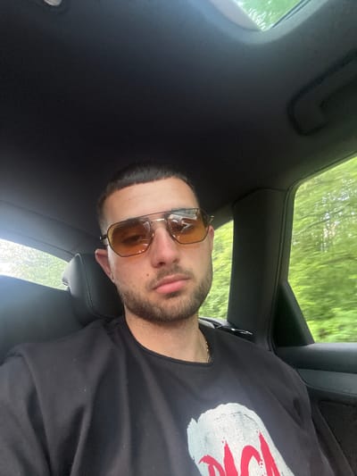 A man wearing sunglasses and a black t-shirt with a white and red design is in the driver's seat of a car. Trees are visible through the sunroof. The interior of the car is black. The location is likely a road in Rahovtsi, Bulgaria based on previous context.
