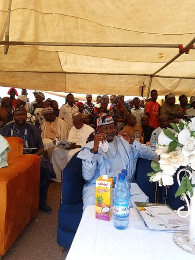 The image shows a gathering of people under a white canopy tent, likely in an outdoor setting. The people are seated on chairs and are dressed in various attire, some wearing traditional clothing. The man in the center of the image, wearing a blue long-sleeved shirt, is wiping his glasses with a tissue. He is seated on a blue chair and in front of him is a table with a white tablecloth. On the table, there is a carton of Olivia D'elite pineapple juice, a bottle of FAR water, and some papers. There are white flowers in a vase to the right of the table. The image was taken in Zum, Nigeria, at latitude 10.116 and longitude 13.047.