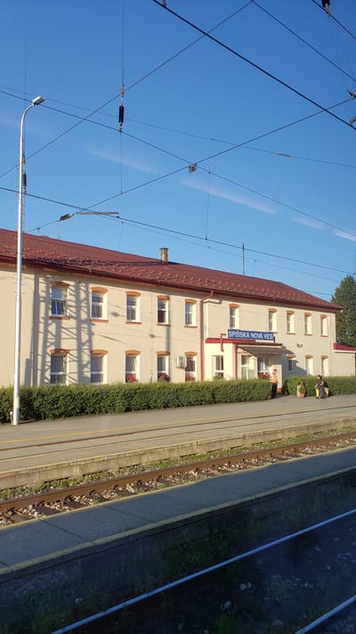 The image shows a train station in Spišská Nová Ves, Slovakia. The station building is a two-story structure with a red tiled roof and cream-colored walls. It has numerous windows and a prominent blue sign that reads "SPIŠSKÁ NOVÁ VES". A bus stop is located in front of the building, and there are railway tracks and a platform in the foreground. Two individuals are visible on the platform, one wearing an orange vest and the other sitting on a bench. The sky is clear and blue, indicating a sunny day.