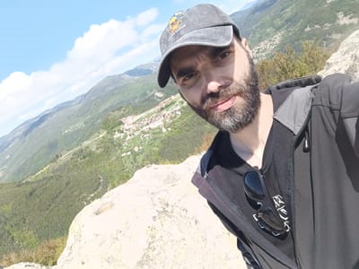 A medium shot captures a man with a beard, wearing a baseball cap, black T-shirt, and dark zip-up jacket, taking a selfie. Sunglasses are attached to his shirt. The background reveals a scenic view of a mountain range and a town below. The location is likely Sini Vrah, Bulgaria, suggested by the landscape and architectural style of the town in the distance. He is standing on a large rock formation. The sky is partially cloudy.