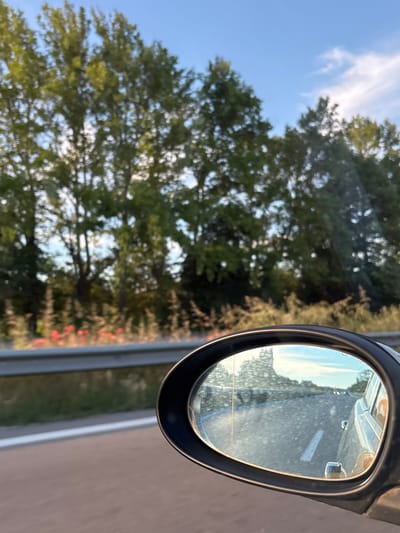 The image shows a reflection in a car's side mirror, depicting a road, trees, and the sky. The reflection shows a road with a white line marking the lane, and a silver car is also visible in the reflection, suggesting the viewer is traveling in a car. Beyond the road, there are trees with green foliage, and the sky is blue with some light clouds. The environment appears to be natural, possibly a rural area or a highway passing through a green landscape. There are no people or specific urban elements visible in the reflection or the surrounding environment. The location is identified as Ignatievo, Bulgaria.