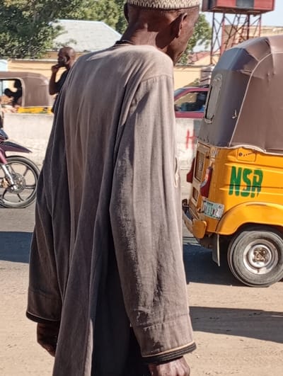 An elderly man in a long, brown robe stands in the street with his back to the viewer. He wears a white cap. A yellow auto rickshaw is partially visible to the right, bearing the letters "NSR" in green. In the background, a motorcycle, a building, and another auto rickshaw can be seen. The scene is bathed in daylight.